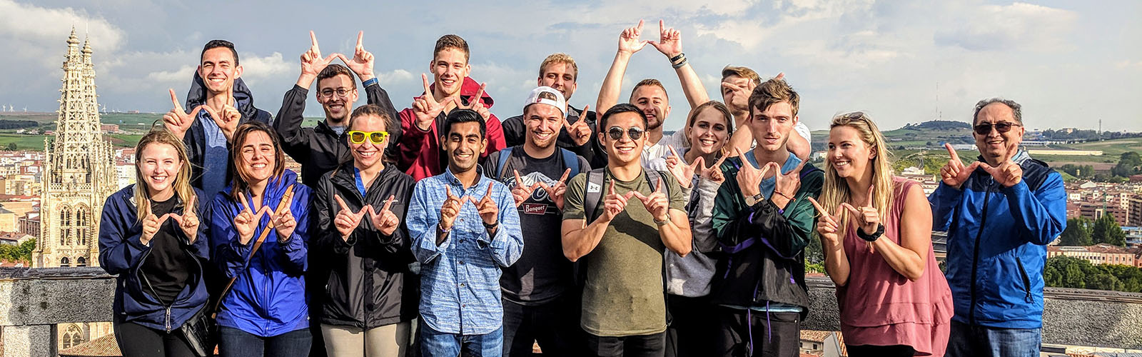 Sixteen individuals posed in two rows, each holding up the letter "W" with their hands. They are at an elevated overlook, with buildings visible behind them.